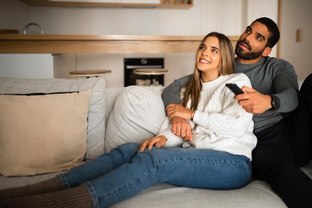 Foto me alegro de que la pareja milenaria multiétnica se abrazo y se siente en el sofá para disfrutar del tiempo libre viendo la televisión junto con el control remoto