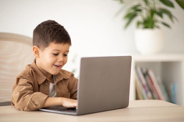 Me alegro de que el pequeño alumno caucásico lindo sorprendido se siente en la mesa escribiendo en la computadora y disfrute de la lección en la habitación
