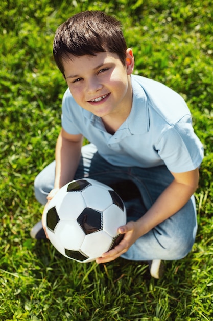 Me gusta el futbol. Muchacho de pelo oscuro alegre sonriendo y sentado con su balón de fútbol