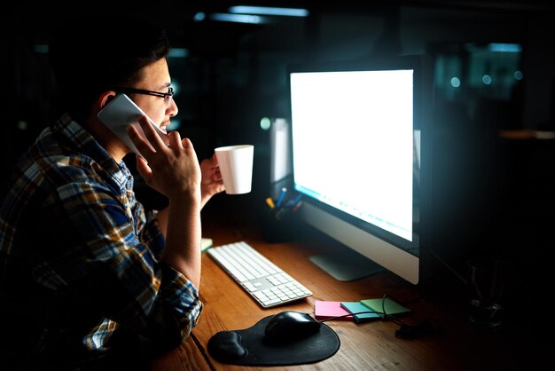 Me iré de la oficina pronto Fotografía de un joven diseñador hablando por teléfono y trabajando en su computadora a altas horas de la noche