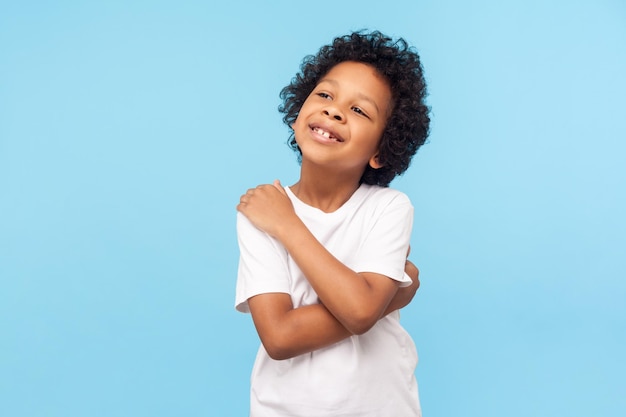 Me quiero a mi misma. Retrato de un adorable niño optimista con rizos abrazándose a sí mismo y sonriendo, un niño egoísta seguro de sí mismo con autoestima positiva. tiro del estudio aislado sobre fondo azul