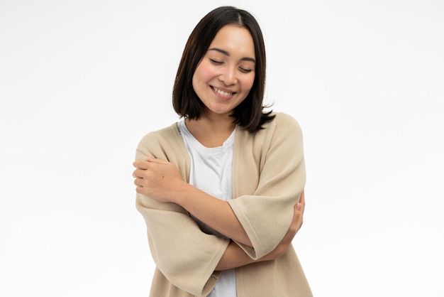 Me quiero a mi misma. Retrato de una chica asiática complacida y satisfecha abrazándose y sonriendo, sintiéndose cómoda y satisfecha, persona egoísta narcisista. Foto de estudio interior fondo blanco.