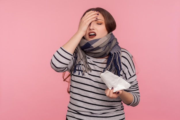 Me siento enfermo Retrato de una mujer malsana envuelta en una cálida bufanda tocando la cabeza dolorida que sufre dolor de cabeza, fiebre y síntomas de gripe sosteniendo una servilleta para estornudar en un estudio interior aislado en un fondo rosado