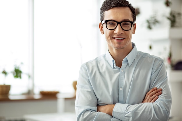 Me siento feliz. Hombre de pelo oscuro alegre atractivo sonriendo y con gafas y con los brazos cruzados