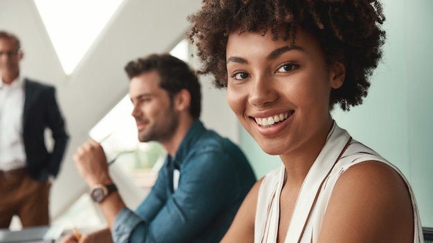 Me siento feliz. Joven afroamericana está mirando a cámara y sonriendo mientras trabaja con colegas en la oficina. Trabajo en equipo. Cita