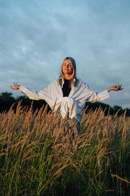 Meaditierende junge blonde Frau, die in der Ferne inmitten eines Weizenfeldes steht
