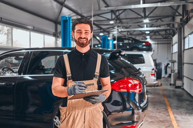 Mecánica en el taller de reparaciones Mecánico joven alegre que escribe algo en sus coches del portapapeles