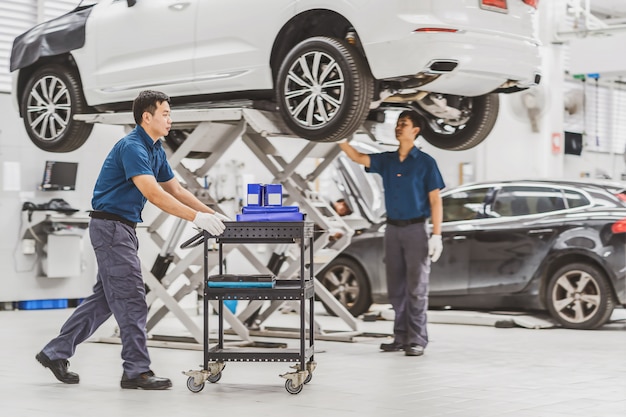 Mecânico asiático empurre o carrinho com o equipamento do carro sobre o colega verificando e reparando o carro