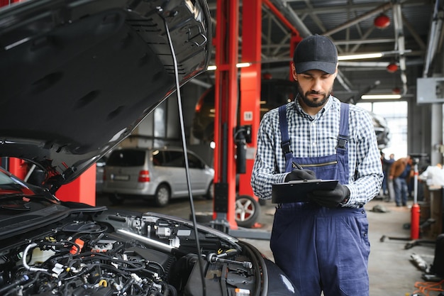 Mecánico de automóviles caucásico profesional con lista de verificación de pie junto al área del motor del vehículo con el capó abierto detectando mal funcionamiento