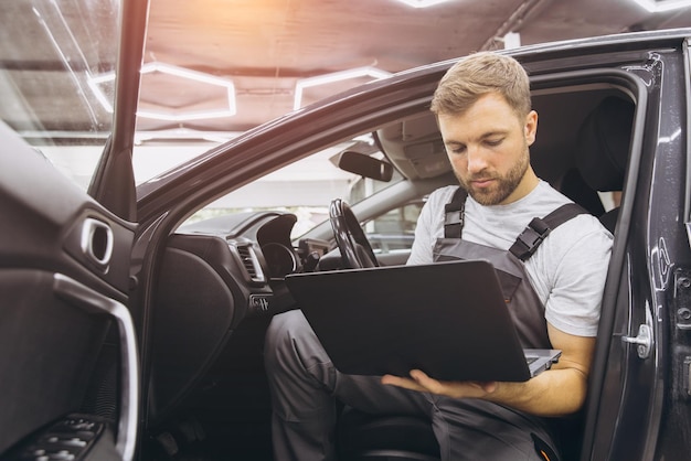 Mecánico de automóviles ejecutando software de diagnóstico en la computadora Mecánico sentado en el coche y trabajando en la computadoras portátiles mientras hace pruebas de diagnóstico de vehículos en el garaje de servicio de automóviles