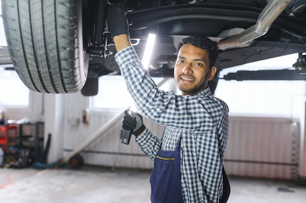 Mecánico de automóviles indio de pie y trabajando en la estación de servicio Especialistas en automóviles examinando el automóvil levantado Reparadores profesionales con uniforme mecánico en color azul