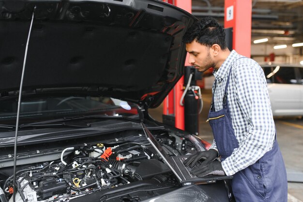 Mecánico de automóviles indio de pie y trabajando en la estación de servicio Especialistas en automóviles examinando el automóvil levantado Reparadores profesionales con uniforme mecánico en color azul