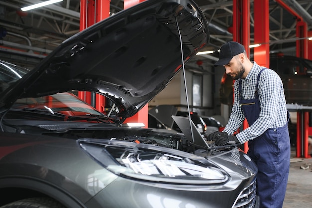 Un mecánico barbudo en uniforme de trabajo está de pie junto a un coche averiado y usando la computadora portátil especial para el mantenimiento del coche Un mecanico en el taller repara el coche