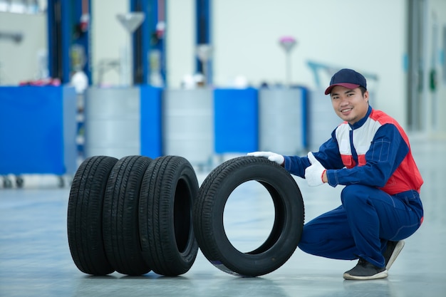 Mecânico bonito asiático de uniforme é sentar e segurar a roda do pneu de carro com o polegar para cima no centro de reparação de automóveis