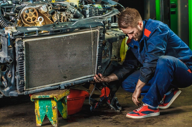 Un mecánico cansado con un traje protector azul está reparando el radiador de un automóvil. Concepto de servicio de reparación.