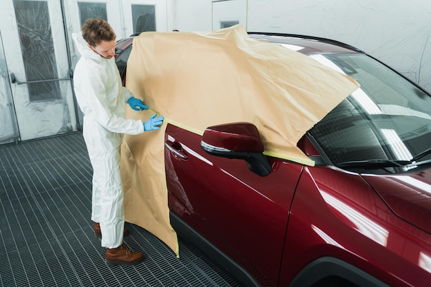 Mecânico cobrindo o carro com papel e fita antes de pintar na cabine de serviço de reparação de automóveis