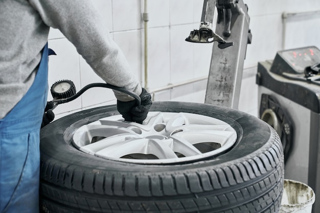 Mecânico de automóveis com suéter cinza e luvas pretas inflando pneu e verificando a pressão do ar com o manômetro na estação de serviço. Conceito de medição e manutenção