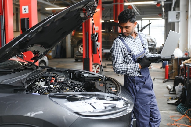 Mecânico de automóveis indiano em pé e trabalhando na estação de serviço Especialistas em automóveis examinando o carro levantado Reparadores profissionais vestindo uniforme mecânico na cor azul