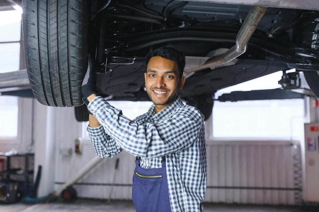 Mecânico de automóveis indiano em pé e trabalhando na estação de serviço Especialistas em automóveis examinando o carro levantado Reparadores profissionais vestindo uniforme mecânico na cor azul