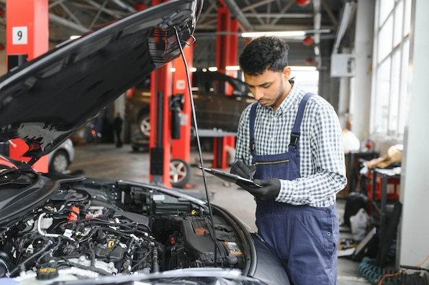 Mecânico de automóveis indiano em pé e trabalhando na estação de serviço Especialistas em automóveis examinando o carro levantado Reparadores profissionais vestindo uniforme mecânico na cor azul