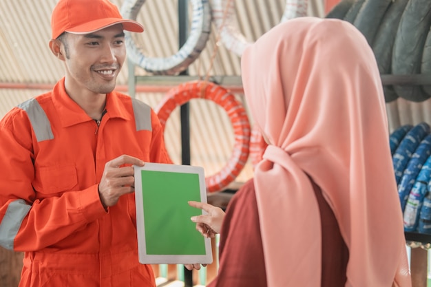 Mecânico em um wearpack usando um tablet digital com um cliente em um suporte para pneus