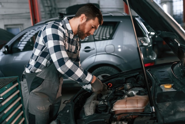 Mecânico está usando uma ferramenta para consertar o carro sob o capô Homem de uniforme está trabalhando