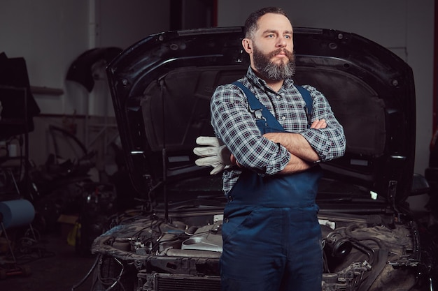 Un mecánico experto barbudo vestido con un uniforme, de pie con los brazos cruzados contra un coche en el garaje.