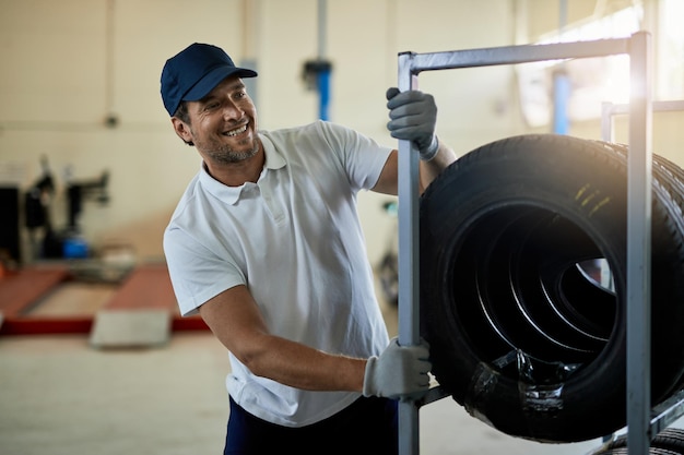 Mecánico feliz empujando un estante con neumáticos de coche nuevos mientras trabaja en el taller de reparación de automóviles