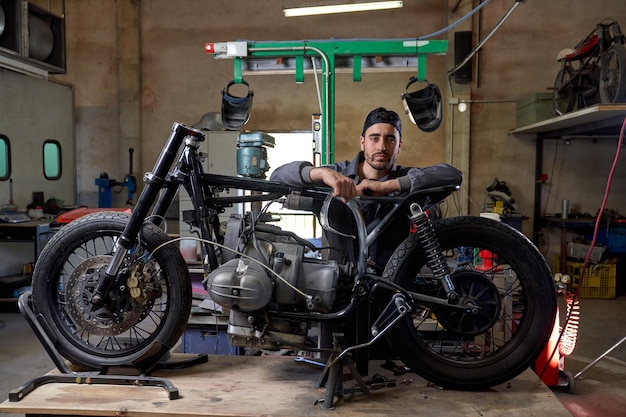 Mecânico masculino barbudo sério, de uniforme, apoiando-se em uma moto desmontada e olhando para a câmera em uma oficina pobre