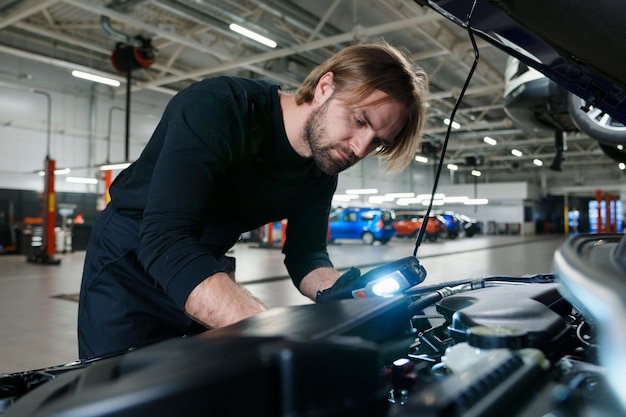 Un mecánico masculino uniformado con una linterna realiza diagnósticos bajo el capó del automóvil Servicio de automóviles moderno y tecnológico