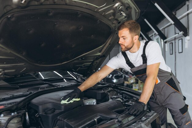 Mecánico en mono limpiando un motor de coche con un paño en un taller de reparación de automóviles