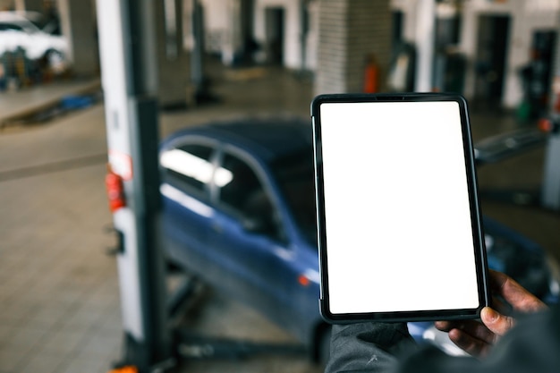 Mecânico segurando um tablet com uma tela em branco em uma oficina de reparo de carros Um carro azul com um capô aberto está no fundo e a oficina tem elevadores de carros e ferramentas visíveis
