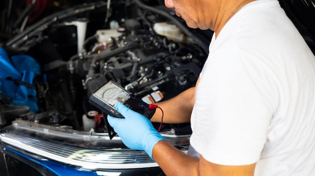 El mecánico senior usa un voltímetro multímetro para verificar el nivel de voltaje en la batería del automóvil en el garaje de servicio y mantenimiento del automóvil