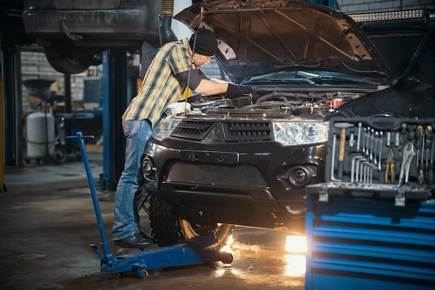 Mecánico de servicio de reparación de automóviles parado junto al automóvil con el capó abierto y trabajando con una herramienta