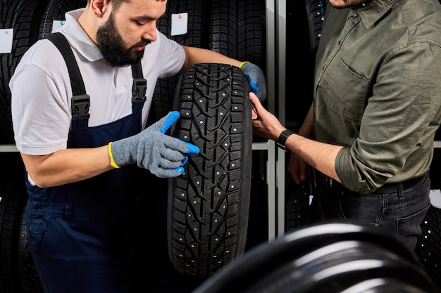 Mecánico sosteniendo un neumático y mostrando los neumáticos de las ruedas en el servicio de reparación de automóviles y la tienda de la tienda de automóviles, joven barbudo en uniforme que trabaja en el taller de reparación de automóviles