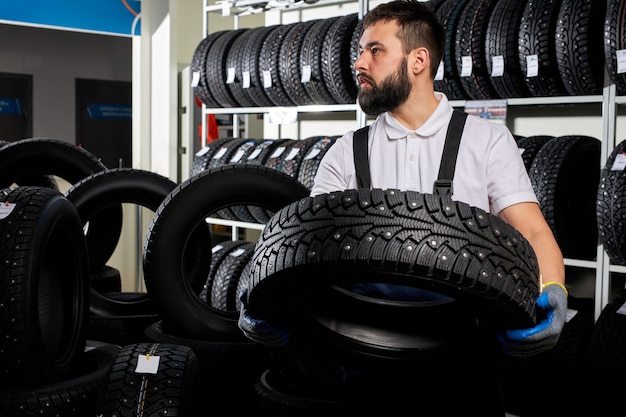 Mecánico sosteniendo un neumático y mostrando los neumáticos de las ruedas en el servicio de reparación de automóviles y la tienda de la tienda de automóviles, joven barbudo en uniforme que trabaja en el taller de reparación de automóviles