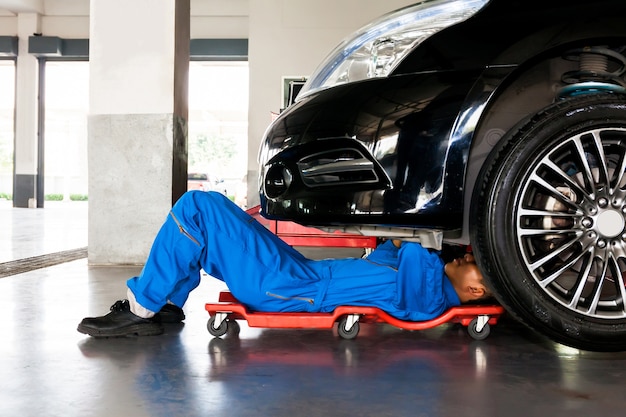 Mecánico en uniforme azul acostado y trabajando bajo el auto en el garaje de servicio de auto