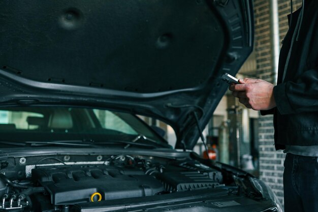 Mecánico usando un teléfono inteligente mientras inspecciona un motor bajo el capó abierto de un automóvil en un garaje profesional Tecnología moderna en diagnóstico y reparación de automóviles