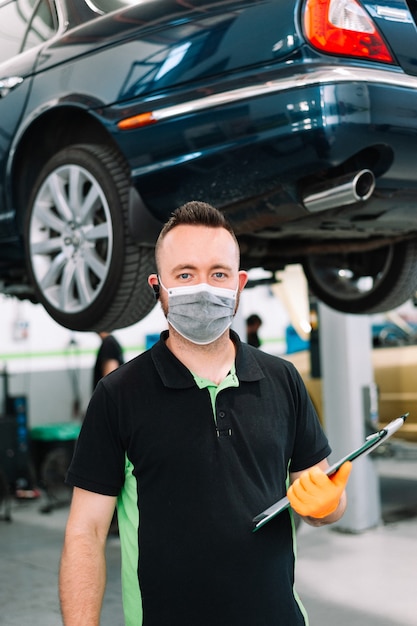 Mecánico vistiendo mascarilla quirúrgica sosteniendo un portapapeles trabajando en el servicio de reparación de garaje