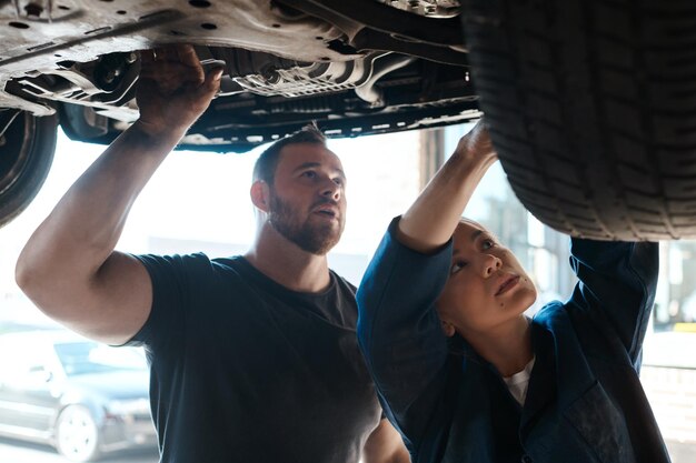 Mechaniker in der Werkstatt und Arbeit an dem Fahrzeugrad mit Ausbildung des Instandhaltungsingenieurs und Teamarbeit am Arbeitsplatz Reparaturwerkstatt Handel und Mentor mit einer Frau Überprüfung unter dem Motor mit Techniker und Auto-Service