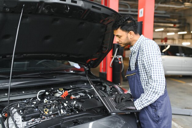 Mechaniker Mechaniker-Manager-Arbeiter mit einem Laptop-Computer, der das Auto in der Werkstatt im Auto-Reparatur-Service-Center überprüft. Junger Mann des Ingenieurs, der sich die Details des Inspektionsfahrzeugs unter der Motorhaube ansieht