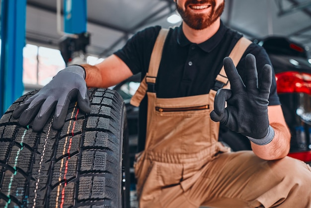 Mechaniker zeigt OK-Geste, hält Autoreifen und lächelt in der Autowerkstatt Zugeschnittenes Bild