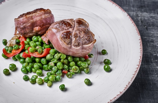 Medallones de ternera envueltos en tocino, servir con guisantes y en un plato sobre fondo de madera.