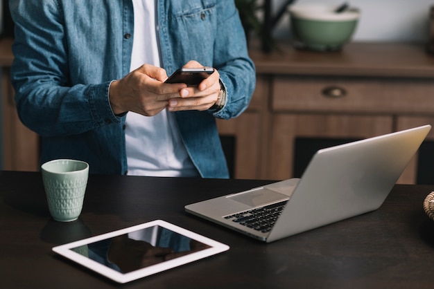 Mediados de sección del hombre joven que usa smartphone con la computadora portátil; tableta digital y taza de café en el mostrador de la cocina