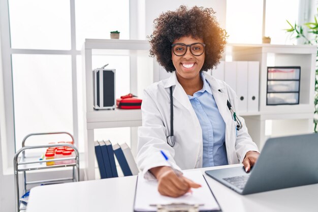 Médica afro-americana usando laptop escrevendo na área de transferência na clínica