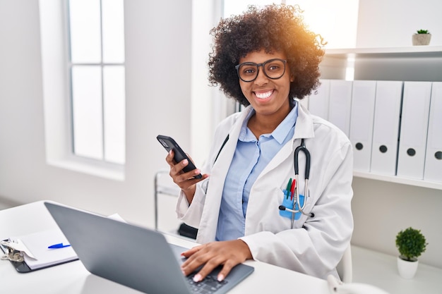 Médica afro-americana usando smartphone e laptop trabalhando na clínica