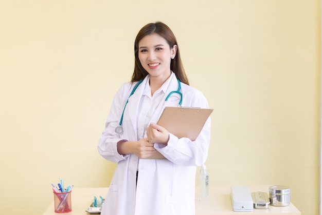 Médica asiática com um jaleco branco se levanta e segura uma prancheta na mão no escritório de trabalho