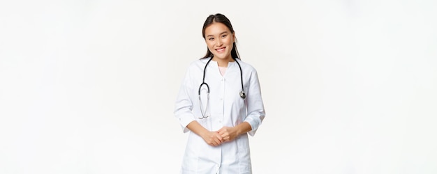 Médica asiática sorridente vestindo roupão médico e estetoscópio parecendo agradável para o paciente em pé