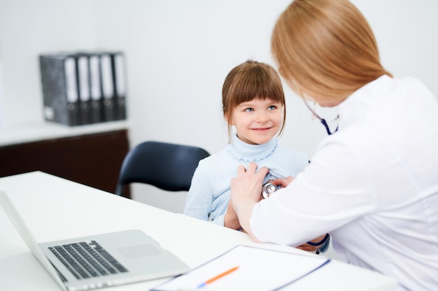 Médica caucasiana examinando paciente garotinha por estetoscópio no escritório