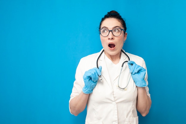 Médica chocada de uniforme usa estetoscópio e escuta em fundo azul